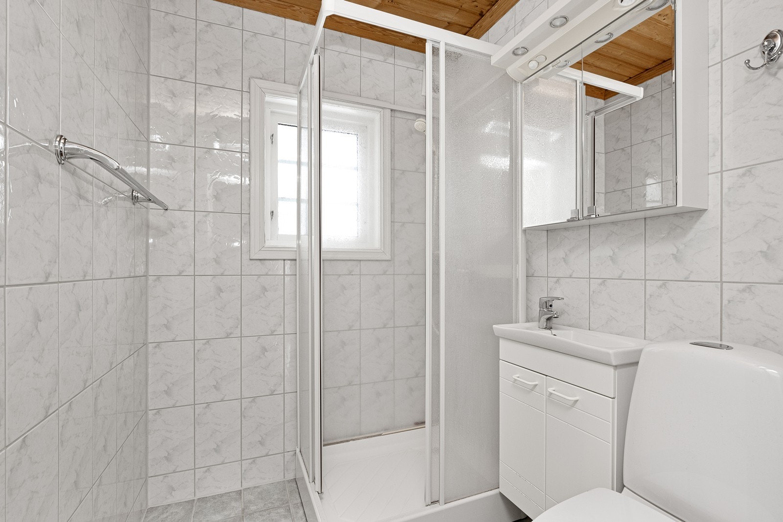 Flislagte overflater på vegger og gulv gir et robust bad, med dusjkabinett plassert i hjørnet. Badet ventileres via elektrisk vifte, og det er lagt vannbåren varme i gulvet for behagelig oppvarming. Galleribilde