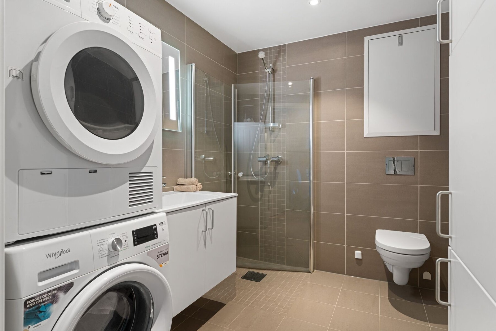 Badet er utstyrt med behagelig varme i gulv, opplegg for vaskemaskin, dusjhjørne, vegghengt toalett og servant med underskap. Rikelig av oppbevaringmuligheter i ekstra skap på vegg. Galleribilde