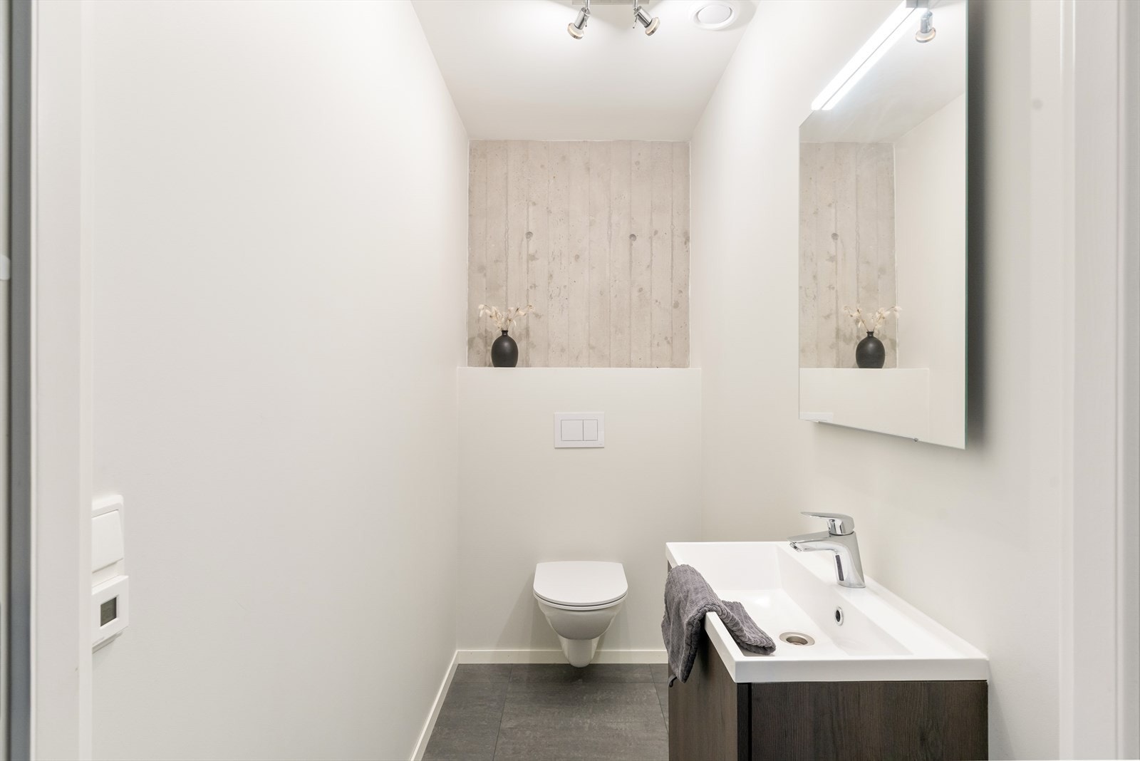 Gjestetoalett med fliser på gulv, vegghengt toalett og vask i seksjon. Betongvegg i bakkant. Varmekabler i gulv Galleribilde