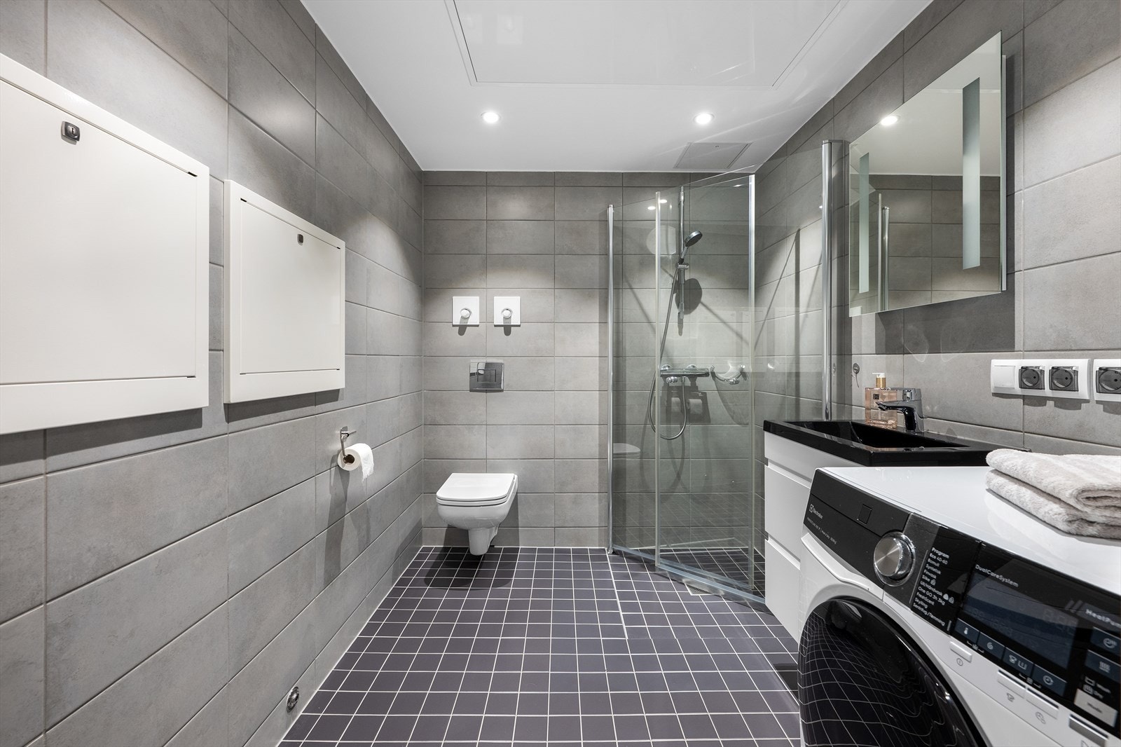 Badet er helfliset og innredet med innsvingbare glassdører i dushjørnet, vegghengt toalett og baderomsinnredning med heldekkende servant og speil på vegg. Det er opplegg til vaskemaskin. Galleribilde