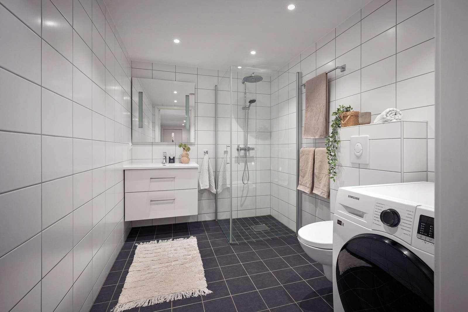 Rommet er innredet med vegghengt toalett, servantinnredning, dusjhjørne med glassvegger samt opplegg for vaskemaskin og tørketrommel. Galleribilde