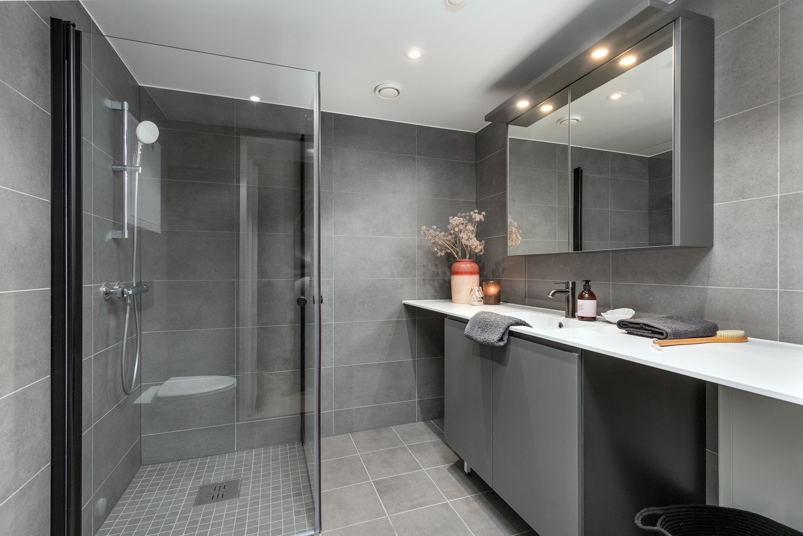 Delikat flislagt bad med vegghengt toalett, dusjsone med innfellbare dører samt servant med skapinnredning. Galleribilde