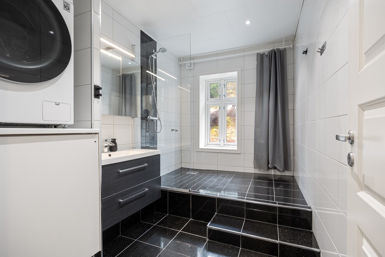 Flislagt baderom med dusj, servant med baderomsinnredning og opplegg til vaskemaskin Galleribilde