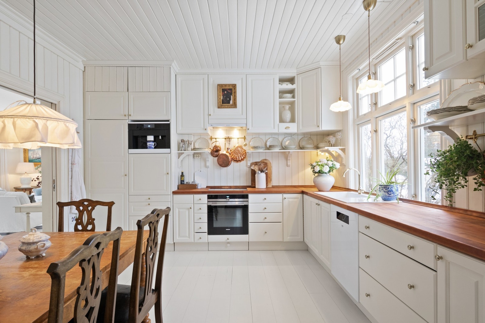 Kjøkkenet er oppført i en funksjonell L-form som gir god arbeidsflyt og rikelig med skapplass. Innredningen fremstår tidløs, med hvite, profilerte fronter, vinhyller, vitrineskap og benkeplate i heltre. Galleribilde