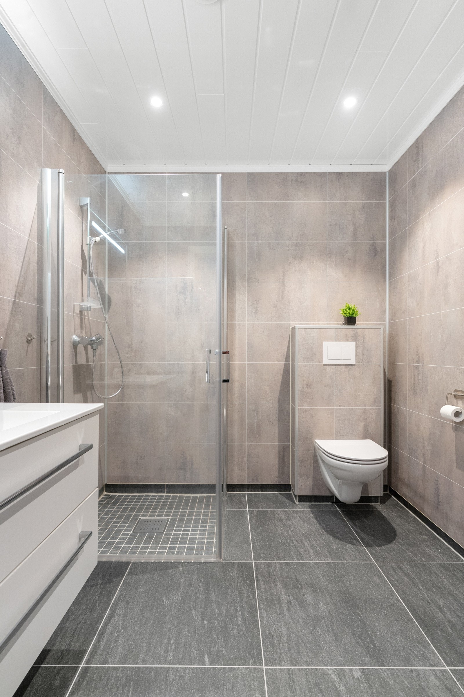 Badet fremstår delikat og funksjonelt, med flislagt gulv, dusjhjørne med glassdører, vegghengt WC og servant med praktisk oppbevaring. Galleribilde