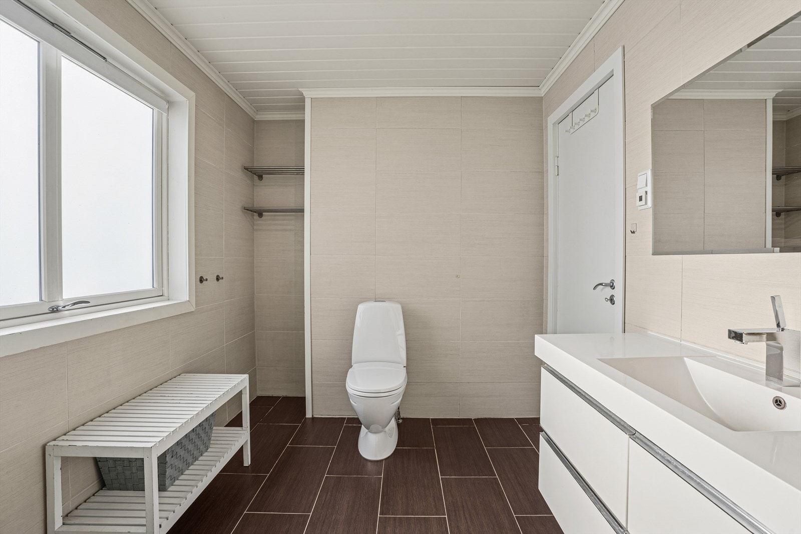 Badet har flislagt gulv med gulvvarme. Fliser på vegger og MDF panel på innvendig tak. Rommet har baderomsinnredning med heldekkende servant og underskap med skuffer, frittstående toalett og dusjkabinett. Galleribilde