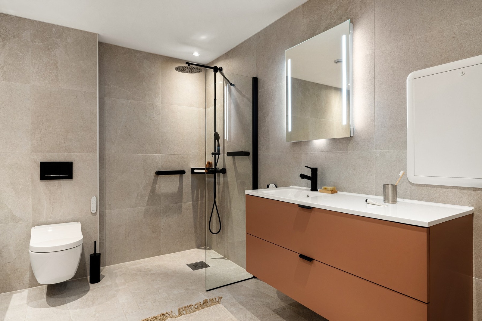 Badet er innredet med vegghengt spyletoalett, dusjhjørne med dusjvegg, badekar og baderomsmøblement med speil og belysning. Galleribilde