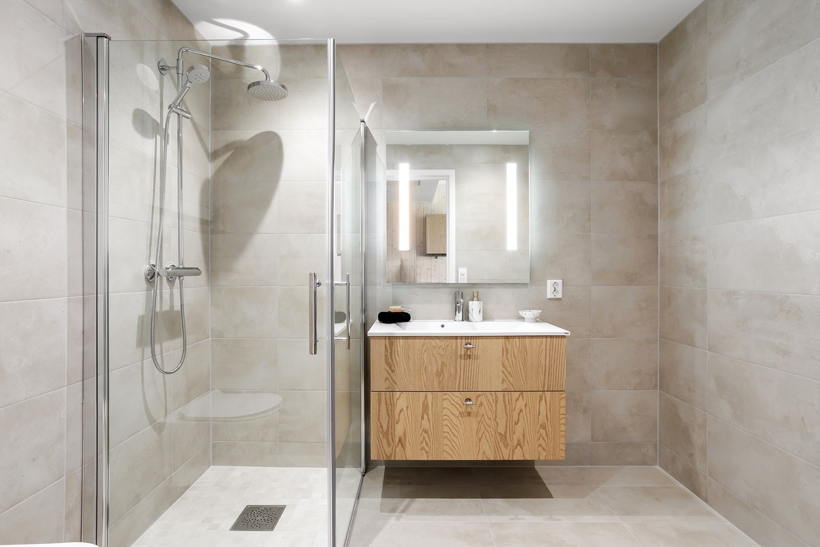 Baderomsinnredning med doble underskap m/kolonialskuffer, heldekkende servantbenk og overliggende speil med integrerte LED-stolper som sammen med integrerte LED downlights i himlingen gir suverent med indirekte lys. Galleribilde