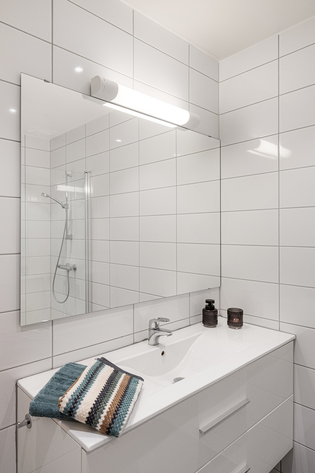 Badet har heldekkende servant over hvit skuffeseksjon og speil i overkant. Videre har badet dusjhjørne med mosaikkfliser og innfellbare dører og vegghengt toalett. Galleribilde