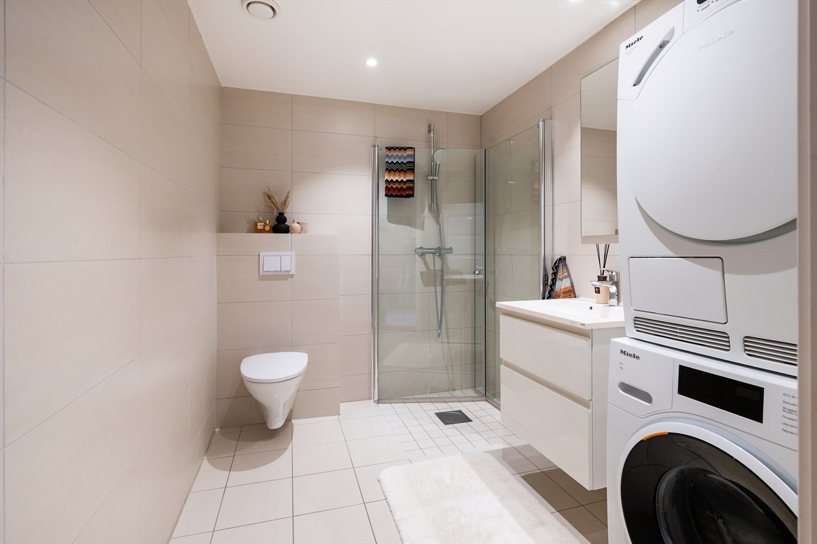 Tidsriktig baderom med vegghengt toalett, heldekkende servant på underskap, speil og belysning. Dusjhjørne med svingbare dører i herdet glass samt opplegg for vaskemaskin og tørketrommel. Galleribilde