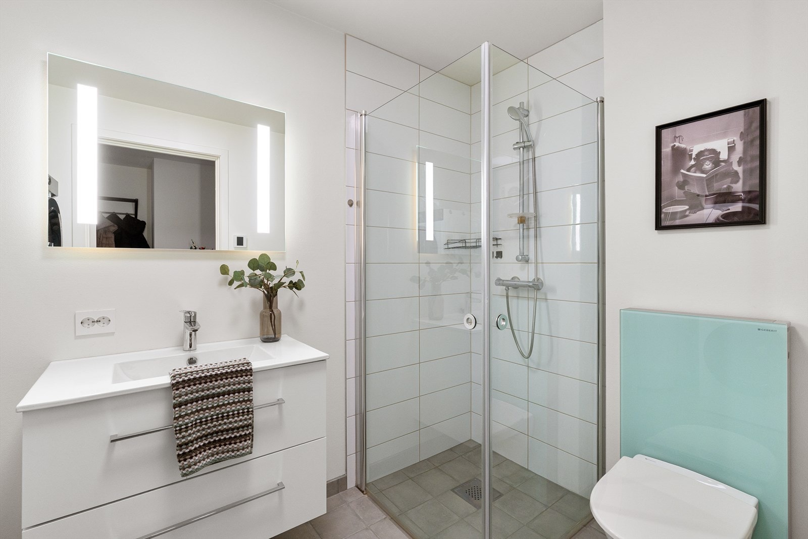 Bad i første etasje med vask i skuffeseksjon. Speil med belysning. Vegghengt toalett. Flislagt gulv og dusjnisje. Galleribilde