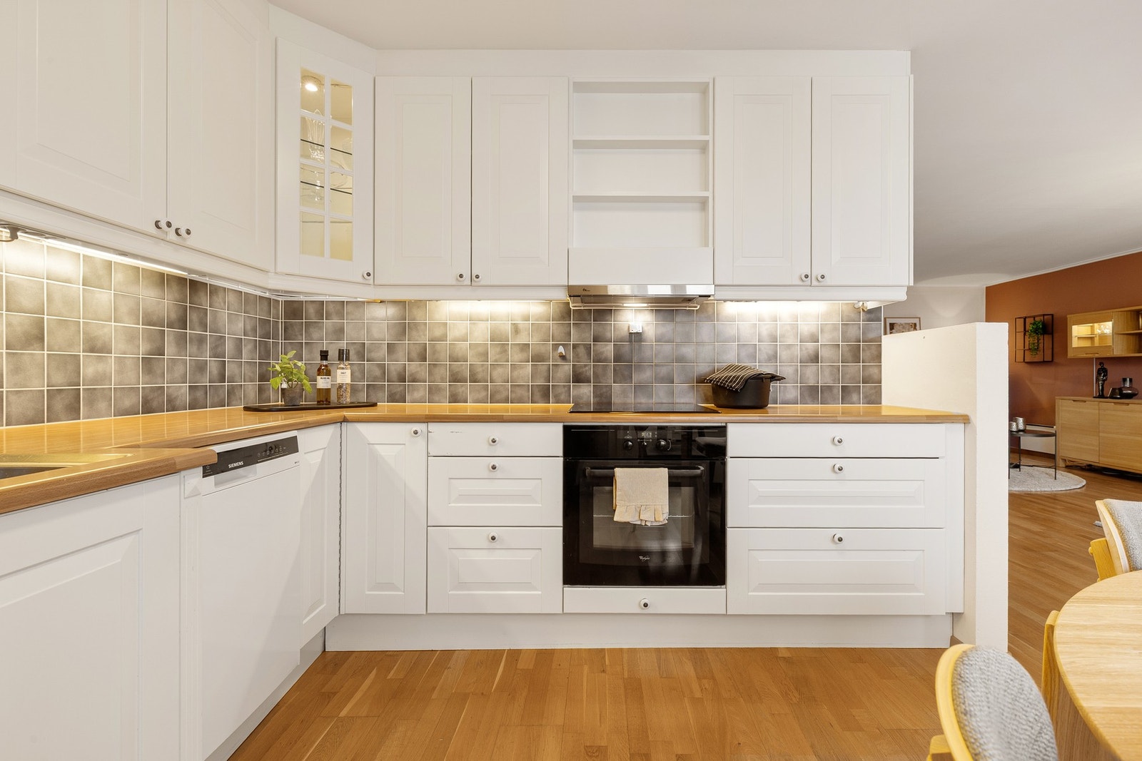 Tidløs innredning med delvis integrerte hvitevarer, fliser over kjøkkenbenk og lys under overskap. Galleribilde