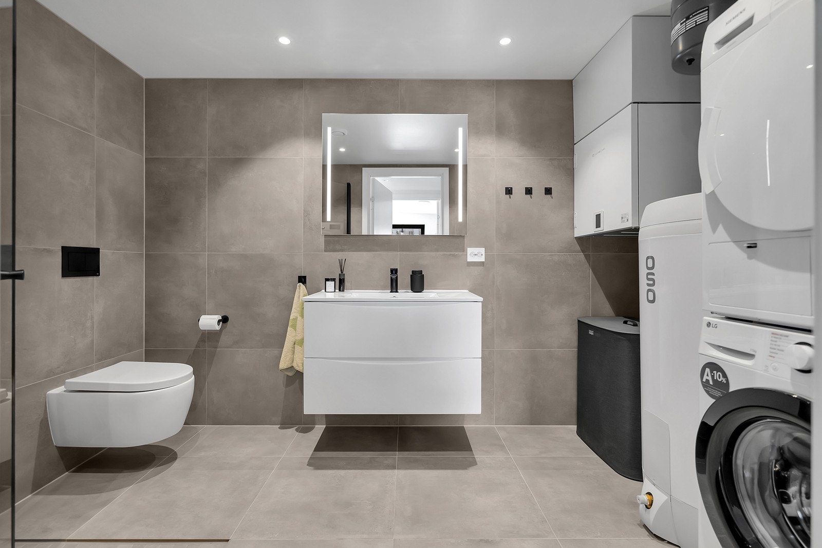 Lekkert og moderne bad med flislagte overflater og varmekabler i gulv. Badet er utstyrt med vegghengt toalett, servantinnredning med skuffer og speil med belysning. Det er også opplegg og plass til vaskemaskin og tørketrommel. Galleribilde