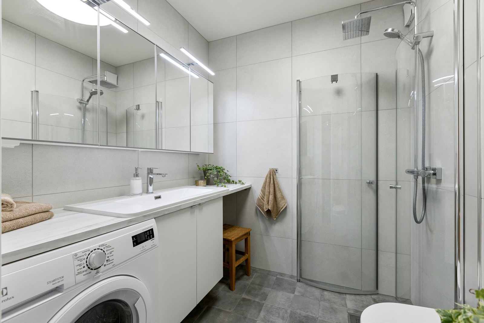 Lyst flislagt baderom med innredningen bestående av servant over underskap, speilskap, dusj med innfellbare dører i glass, samt WC Galleribilde