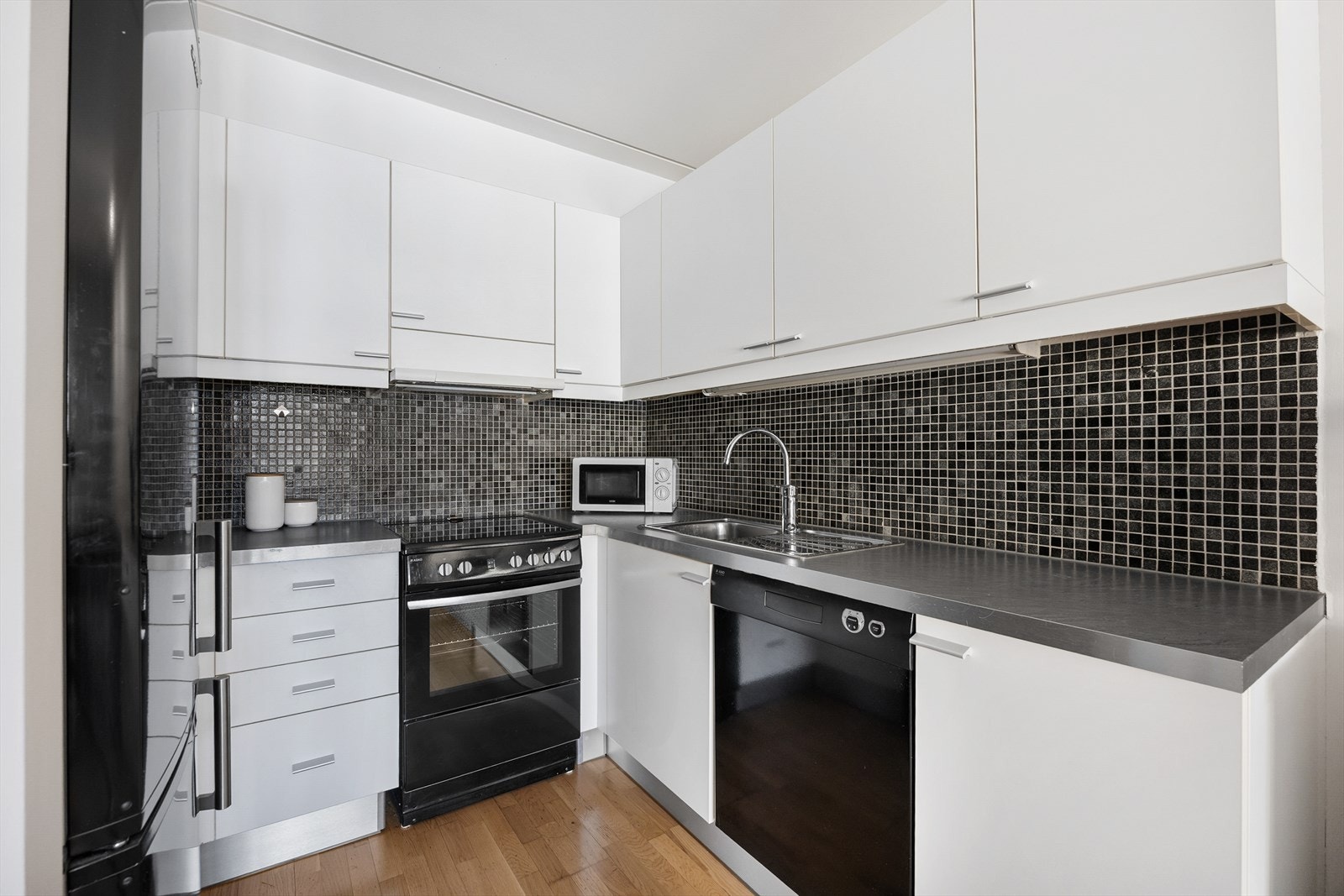 Kjøkkeninnredningen har hvite, glatte fronter og laminert benkeplate. På veggen mellom overskap og benkeplaten er det fliser. Galleribilde