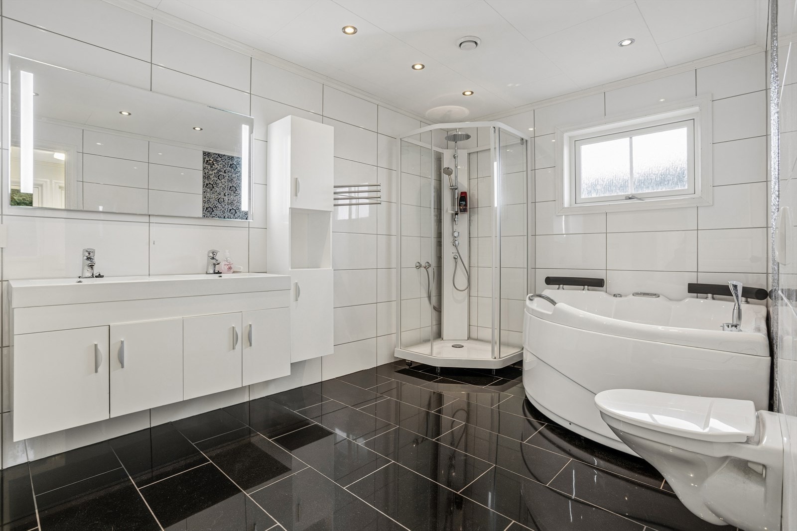 Stort og moderne bad med fliser på gulv og vegger, dobbel servant, høyskap, dusjkabinett, boblebadekar og vegghengt toalett. Galleribilde