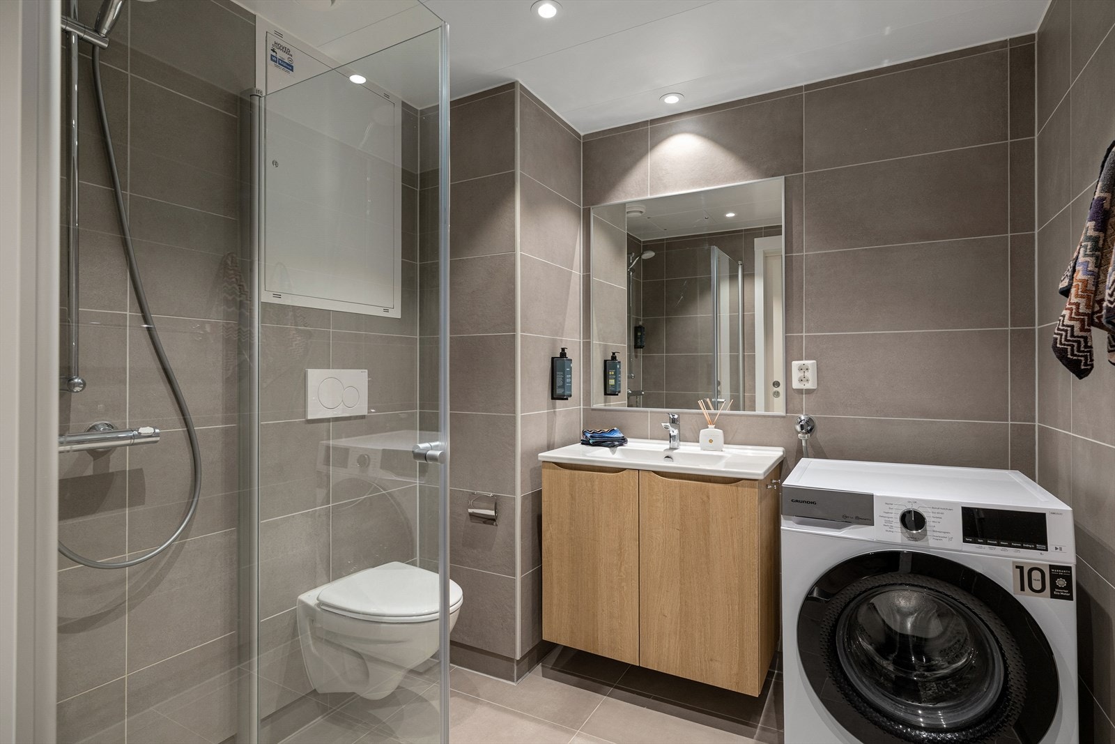 Delikat baderom med servantinnredning, speil, vegghengt toalett og dusjhjørne. Det er opplegg for vaskemaskin på badet Galleribilde