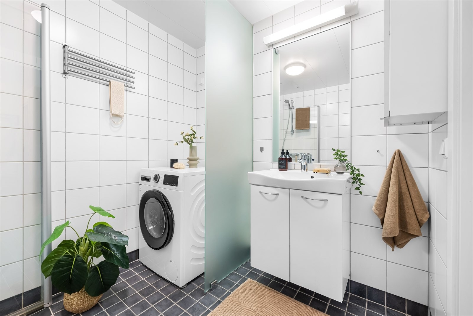 Badet inneholder vegghengt toalett, benkeskap med servant, opplegg for vaskemaskin, fordelerskap til rør i rør vannforsyning og dusjdører i glass. Galleribilde