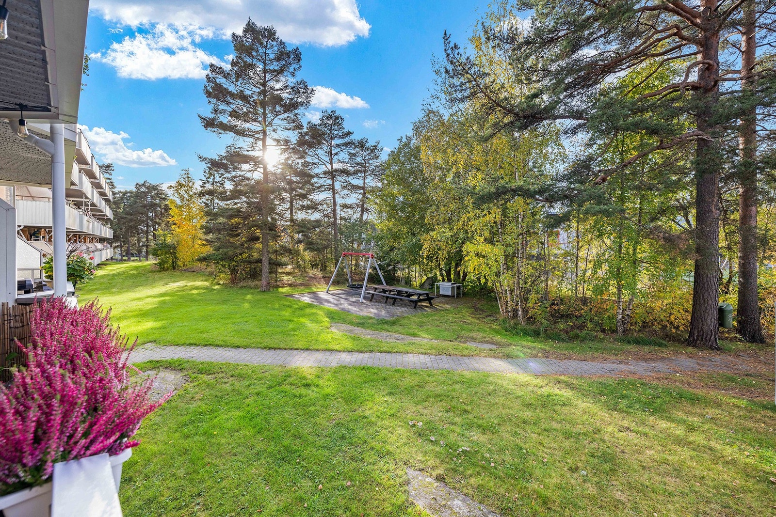 Balkongen vender ut mot fellesareal som er pent opparbeidet med gangstier, grøntarealer, sittegrupper og lekeapparater Galleribilde