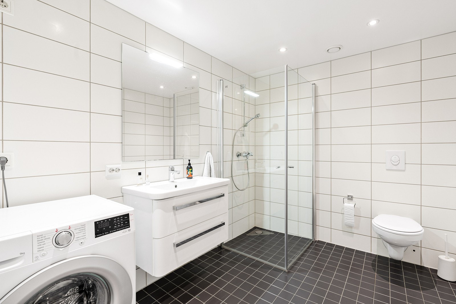 Pent helfliset bad innredet med baderomsinnredning med servant, vegghengt toalett, dusj og opplegg for vaskemaskin. Galleribilde