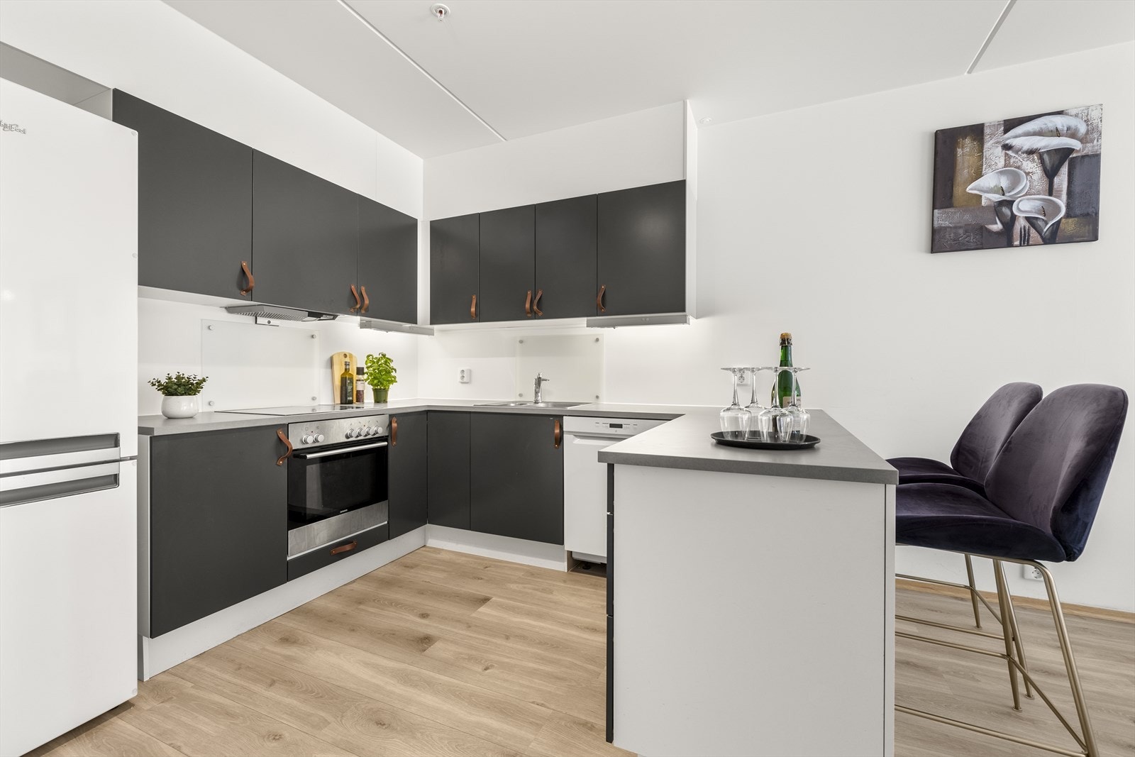 Benkeplaten i lysgrå laminat gir rikelig med arbeidsflate, og glassplater beskytter veggen over vask og koketopp. Galleribilde