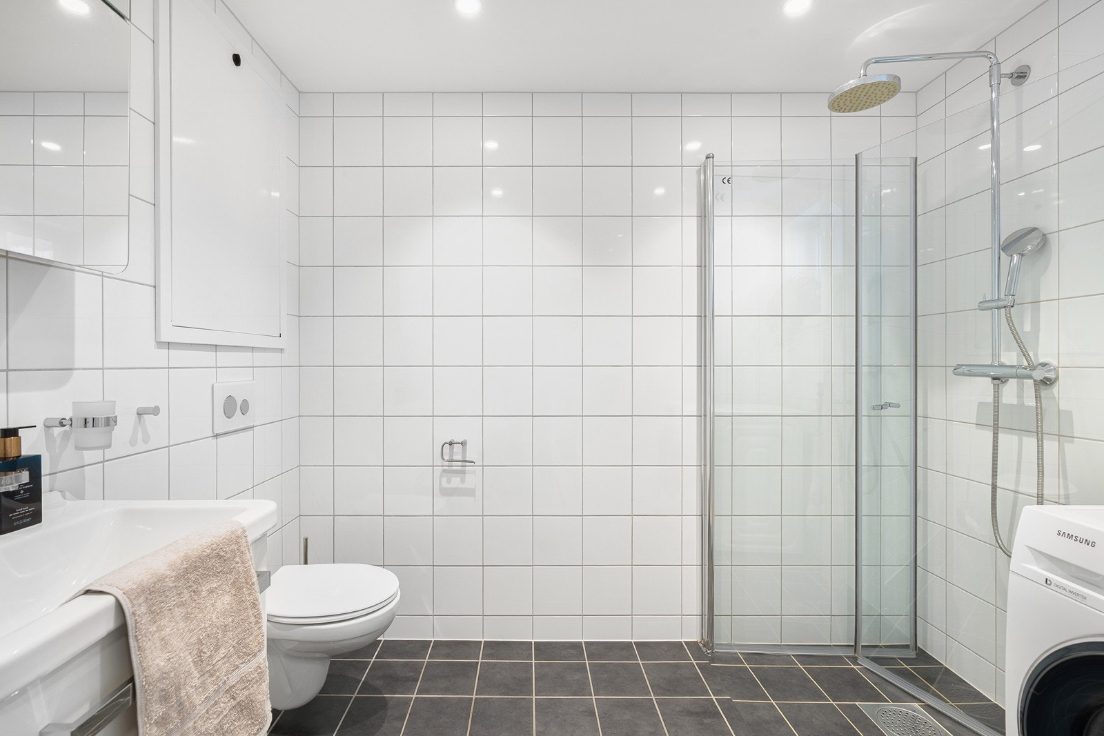 Badet har flislagte overflater, vannbåren gulvvarme og downlights i himling. Rommet er utstyrt med en baderomsinnredning med nedfelt servant, veggmontert toalett og dusjhjørne med innfellbare dusjdører. Galleribilde