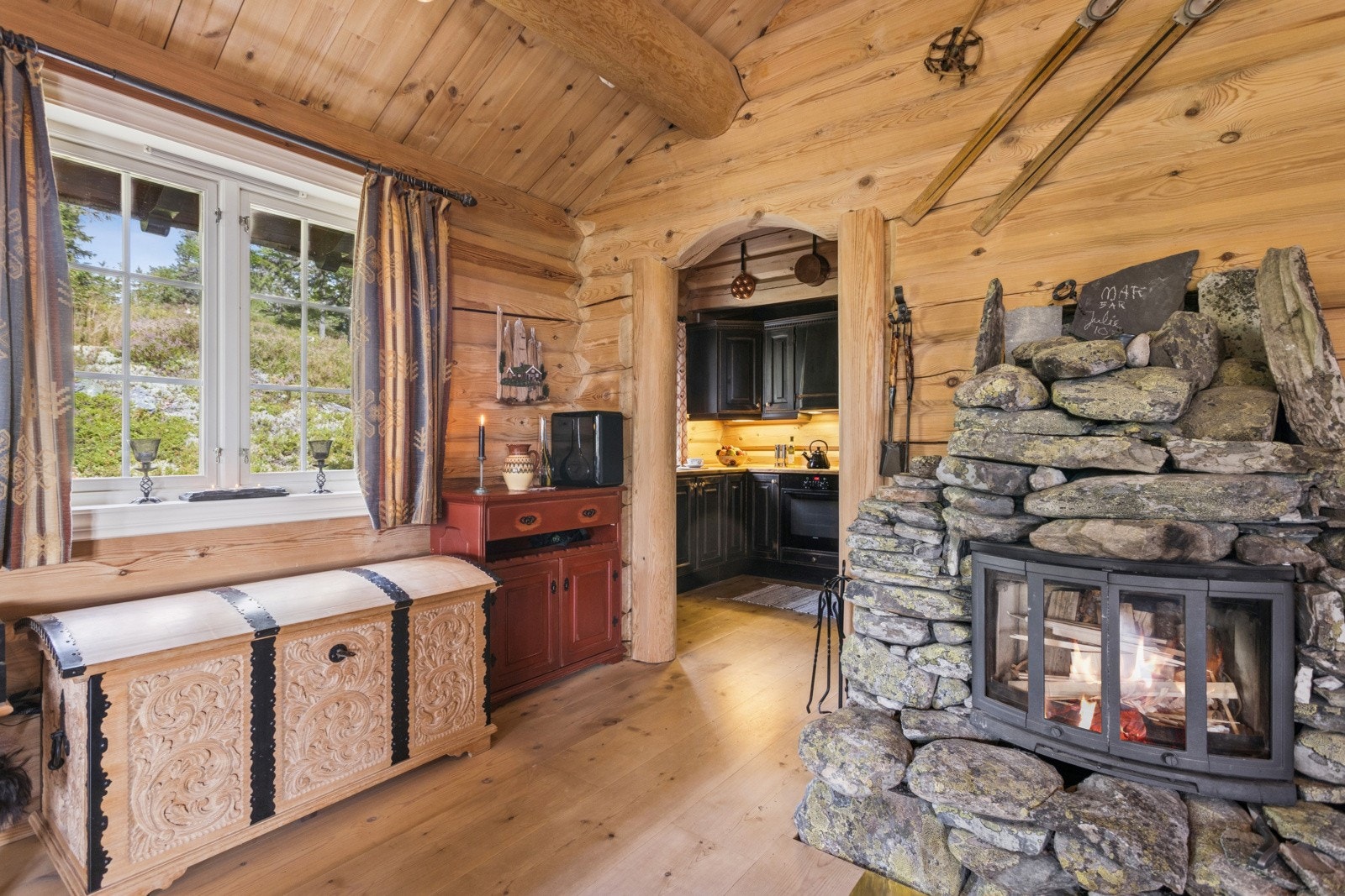 Kjøkkenet ligger godt plassert og har adomst fra stuen. Merk også den stilige peisen - kledd i naturstein, som gir et flott særpreg. Bildet viser også utsikten mot baksiden av hytta med naturtomten som endrer seg i takt med årstidene og gir et fint skue. Galleribilde