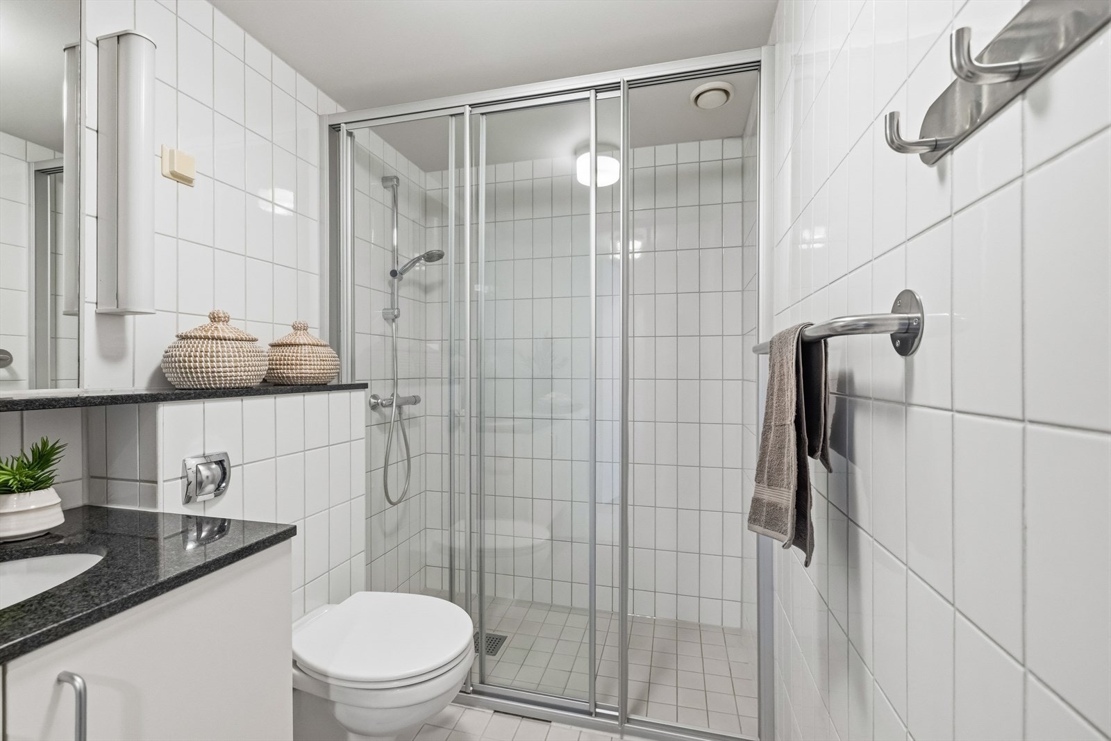 Dusjhjørne med skyvedør, sluk i sone og håndholdt dusjhode samt veggmontert toalett. Galleribilde