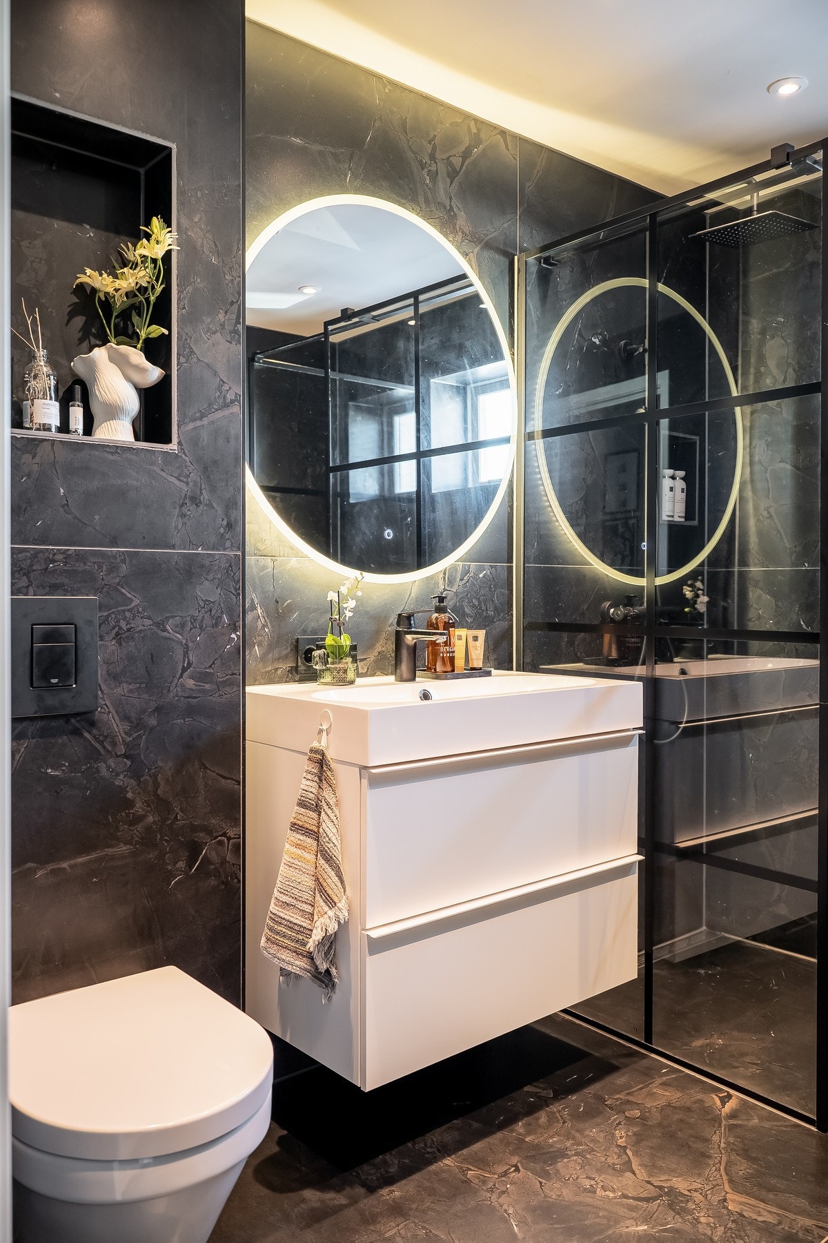 Moderne baderom med dusjhjørne, vegghengt toalett og servant med skuffer. Mørke fliser og speil med lys. Galleribilde