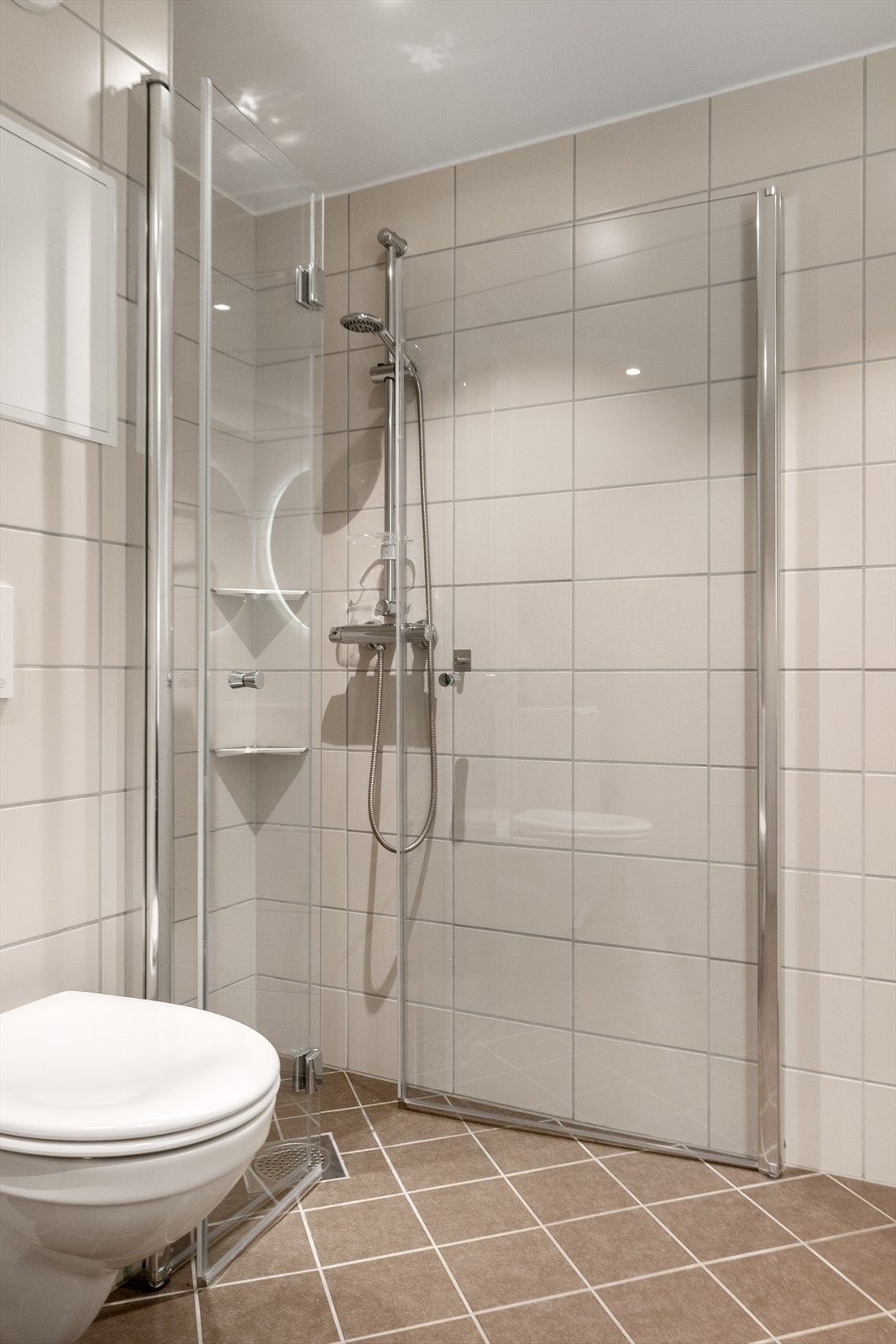 Rommet er innredet med et moderne servantskap med servant, veggmontert toalett og en vegghengt dusj med skjermdører i glass. Galleribilde