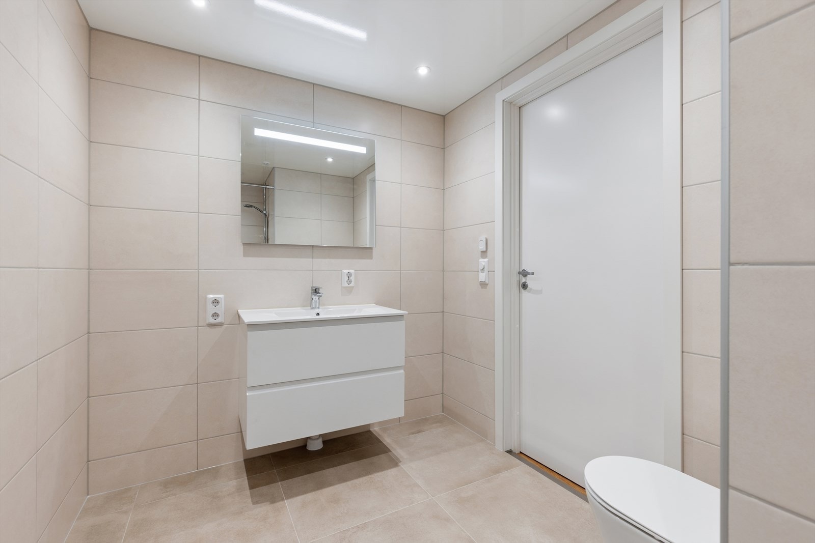 Flislagt baderom med vannbåren varme i gulvet. Badet er utstyrt med baderomsmøbel med servant, dusjnisje og vegghengt toalett. Galleribilde