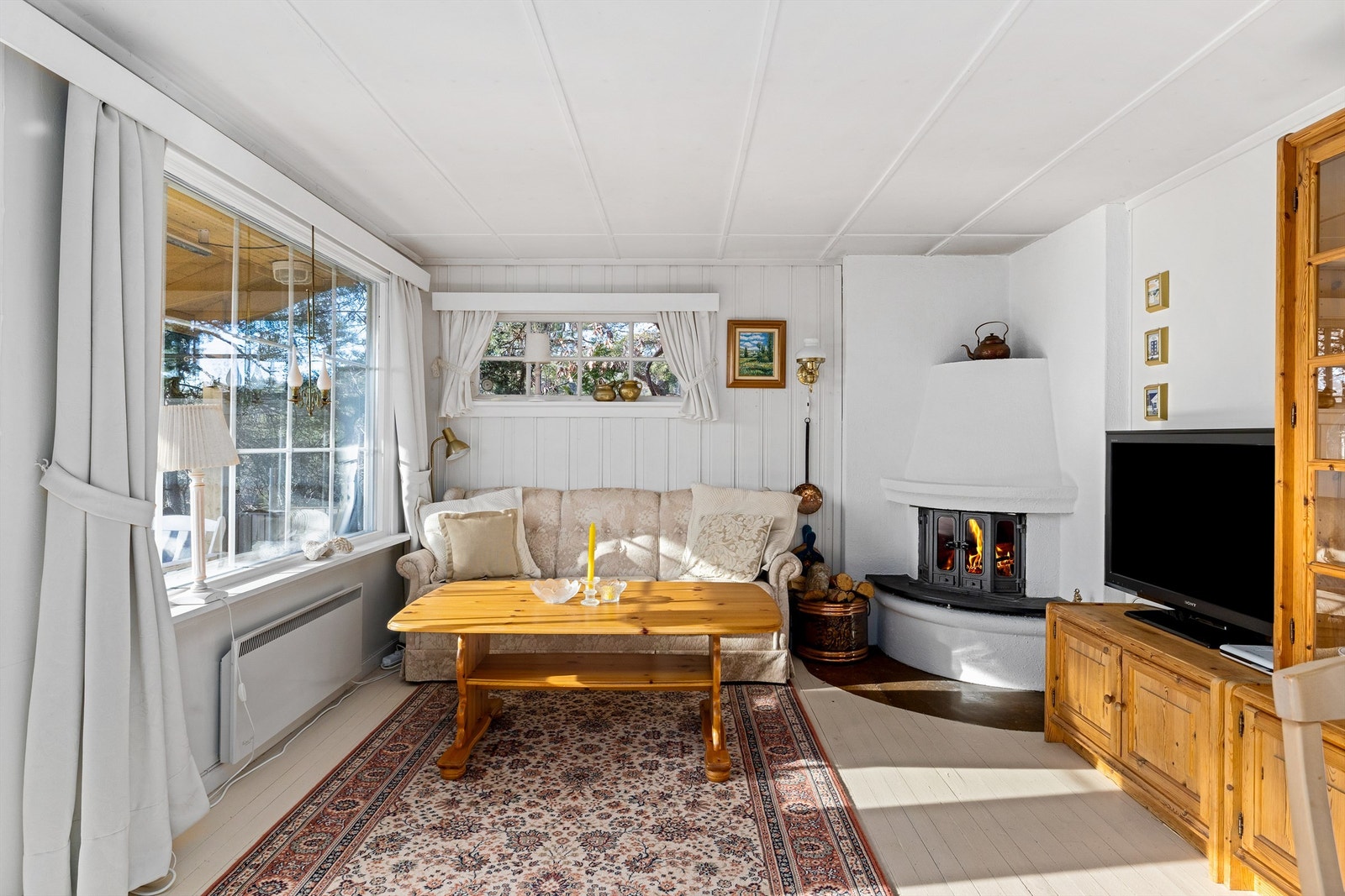 HYGGESTUEN: Peisen er plassert i hjørne slik at flammene kan sees fra hele rommet. Med malte flater og stort vindu mot vest er rommet lyst og trivelig. Galleribilde