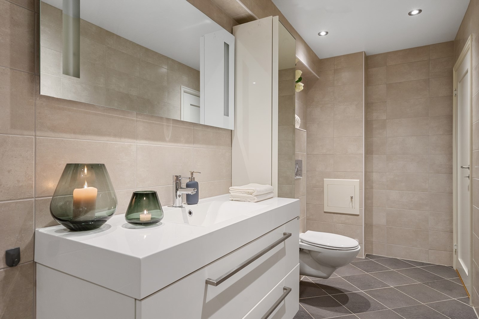 Badet er helfliset i tidløse farger og har gulvvarme og downlights som gir et moderne preg. Det er innredet med stilrene møbler, heldekkende servant, speil med belysning og et praktisk høyskap med speildør. Galleribilde