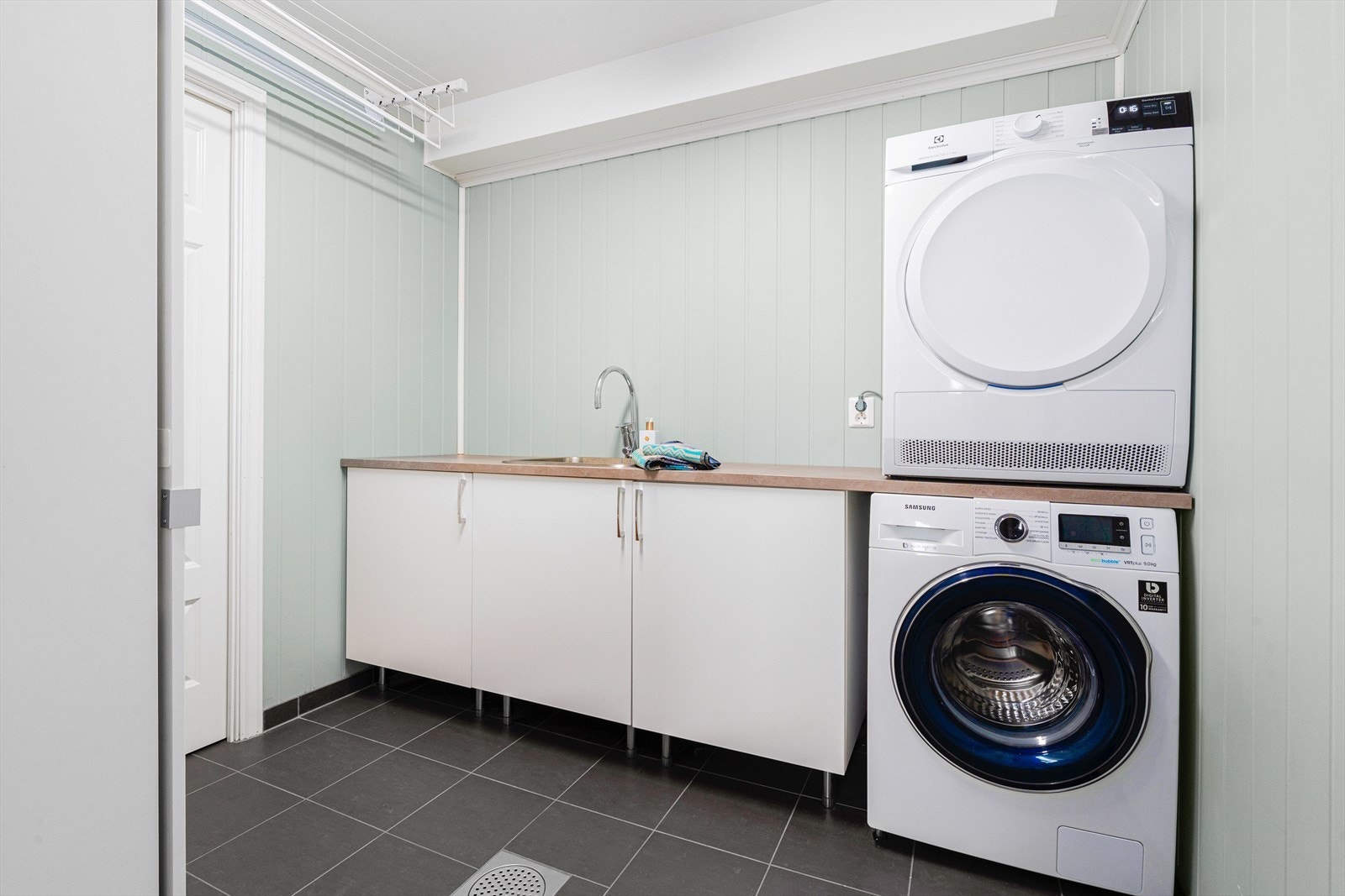 Praktisk vaskerom med plass til diverse oppbevaring. Rommet har benkeplate med skylle kum og det er opplegg for vaskemaskin og tørketrommel. Galleribilde