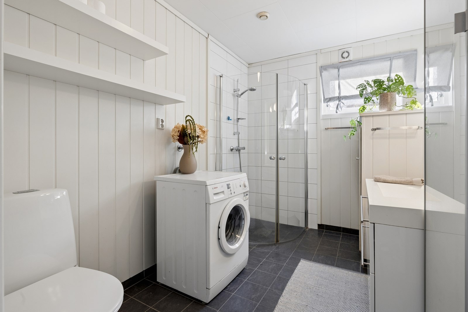 Baderom i hovedetasjen med flislagt gulv, dusjhjørne med svingbare dører, toalett og opplegg til vaskemaskin og tørketrommel. Galleribilde
