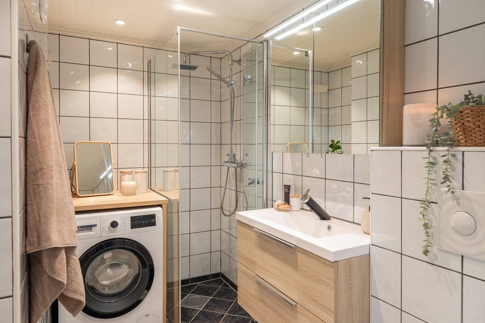 Pent, flislagt baderom med dusjhjørne, servant med underskap, speilskap, vegghengt toalett og opplegg til vaskemaskin. Galleribilde