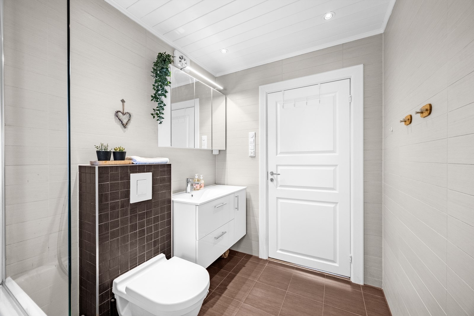 Badet er utstyrt med vegghengt toalett, innmurt badekar og en servantinnredning med skuffer. Praktisk speilskap med belysning på vegg. Galleribilde