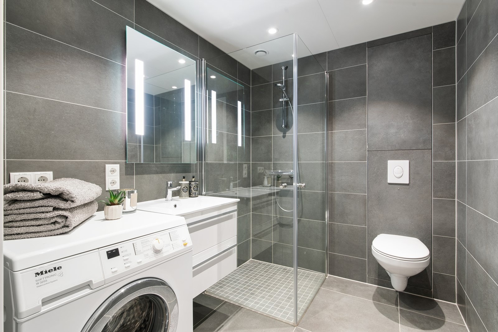 Badet er godt utstyrt med vegghengt toalett, servant og dusjhjørne. I tillegg er det er praktisk oppbevaringsplass under servanten og opplegg for vaskemaskin. Galleribilde