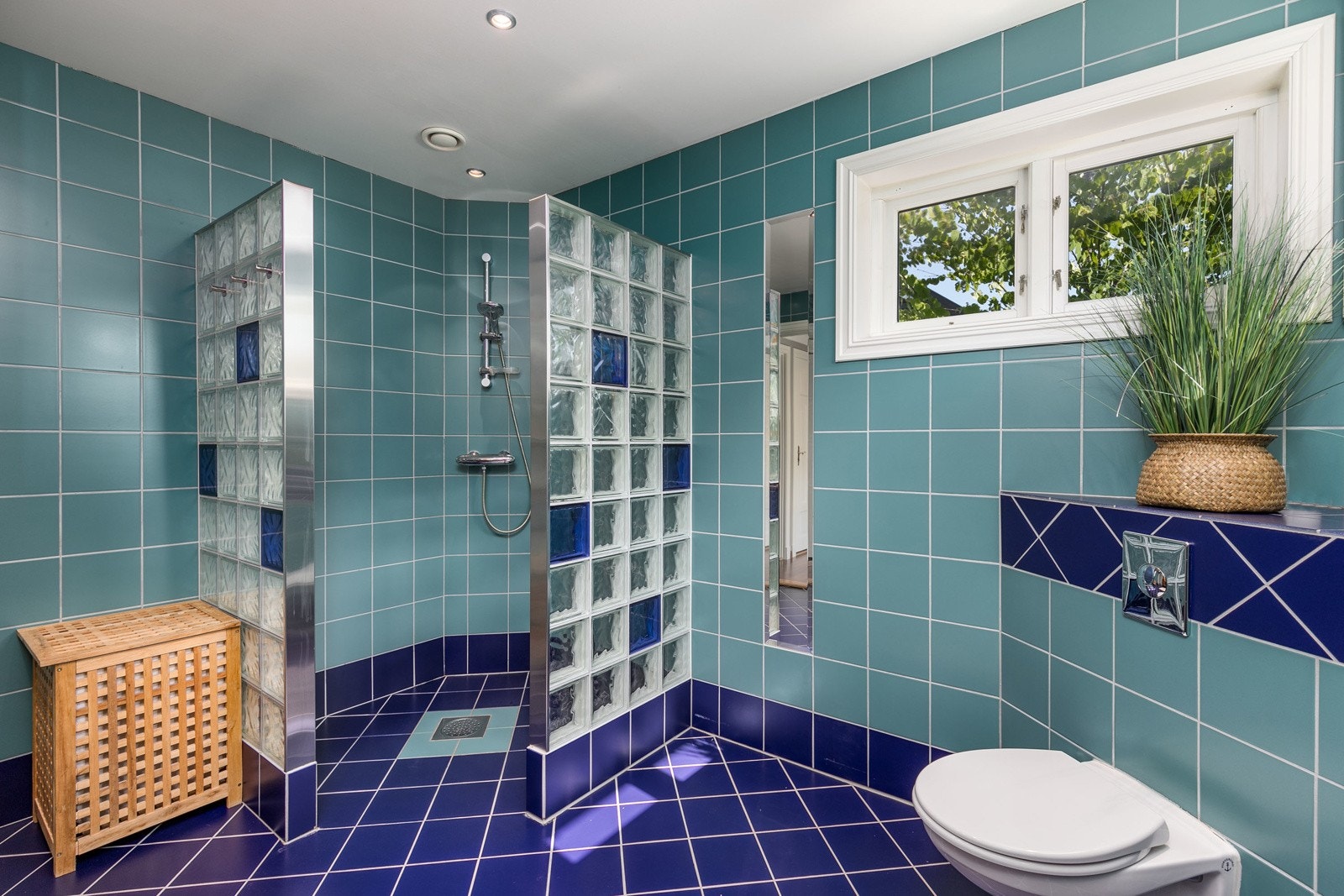 Badet har dusj på gulv i dusjsone med glassbyggersten. Veggfestet klosett med innebygget sisterne. Servanter. Videre har badet benkeskap med glatte fronter, samt overskap. Galleribilde