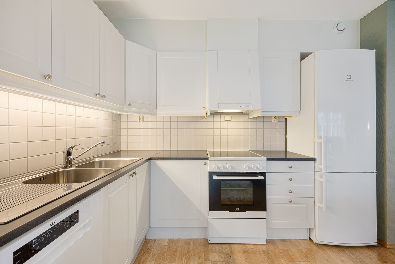 Innredningen har hvite, profilerte fronter og grå laminatbenkeplate med nedfelt kum i stål. Veggene over benken er kledd med hvite fliser, og under overskapene er det montert god arbeidsbelysning. Galleribilde