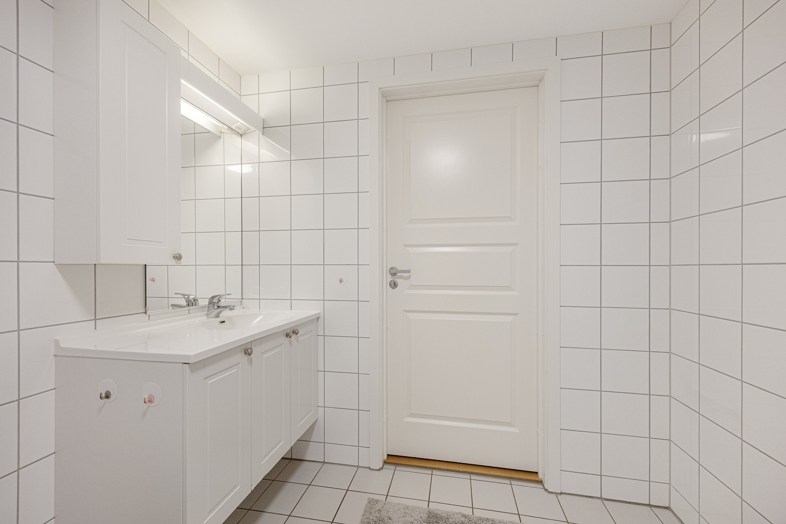 Badet i leiligheten fremstår som velholdt og pent, med hvite fliser på veggene og matchende fliser på gulv med gulvvarme. Innredningen har hvite, profilerte fronter, heldekkende servant, skap og speil. Galleribilde