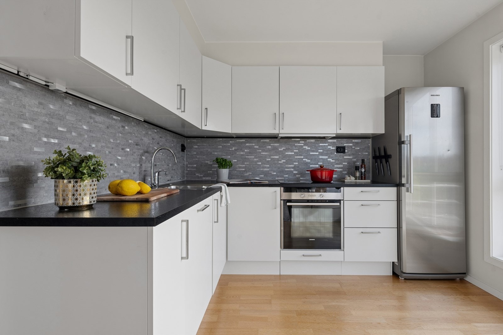 Det moderne kjøkkenet fremstår stilrent og innbydende, med parkett på gulv og malte plater på vegger som skaper en lys og helhetlig atmosfære. Innredningen har slette, moderne fronter og en laminert benkeplate som både er praktisk og lekkert utformet. Galleribilde