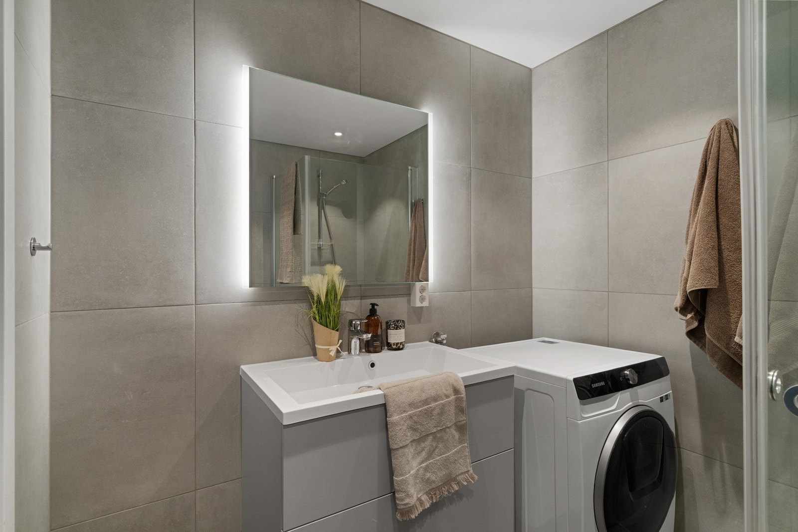Delikat flislagt bad med moderne baderomsinnredning og speil med innfelt lys. Opplegg for vaskemaskin. Galleribilde