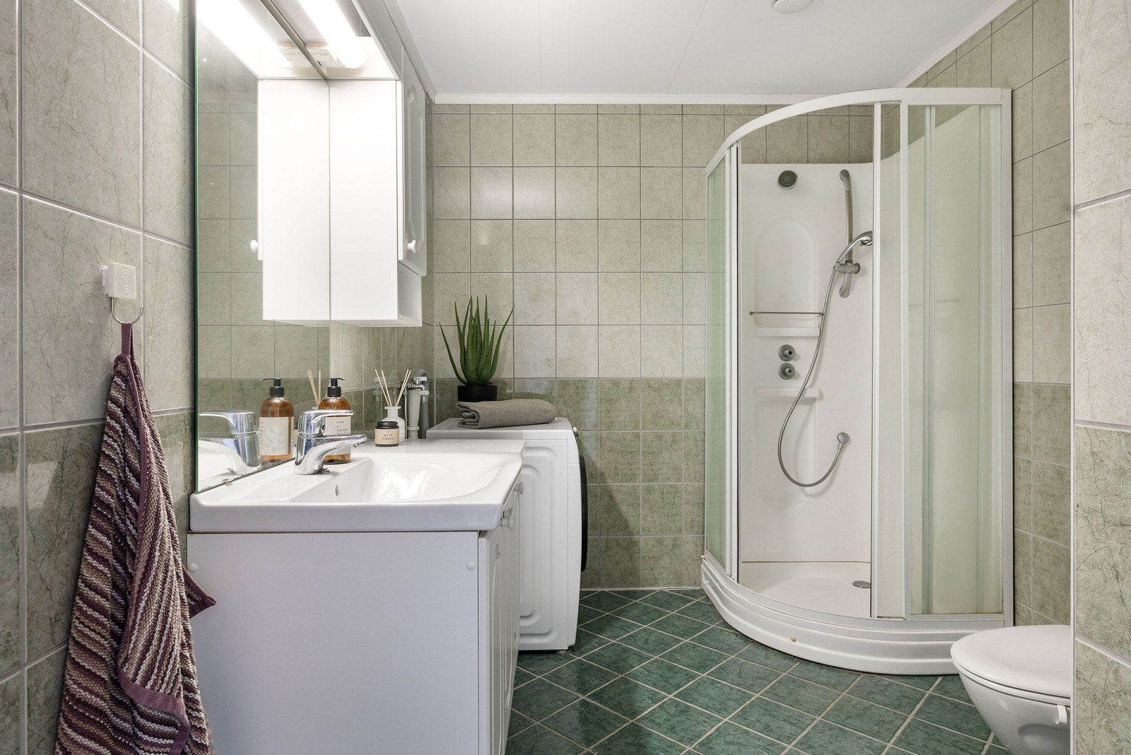 Flislagt bad med opplegg for vaskemaskin. Det er innredet med dusjkabinett, servant i innredning og toalett. Galleribilde