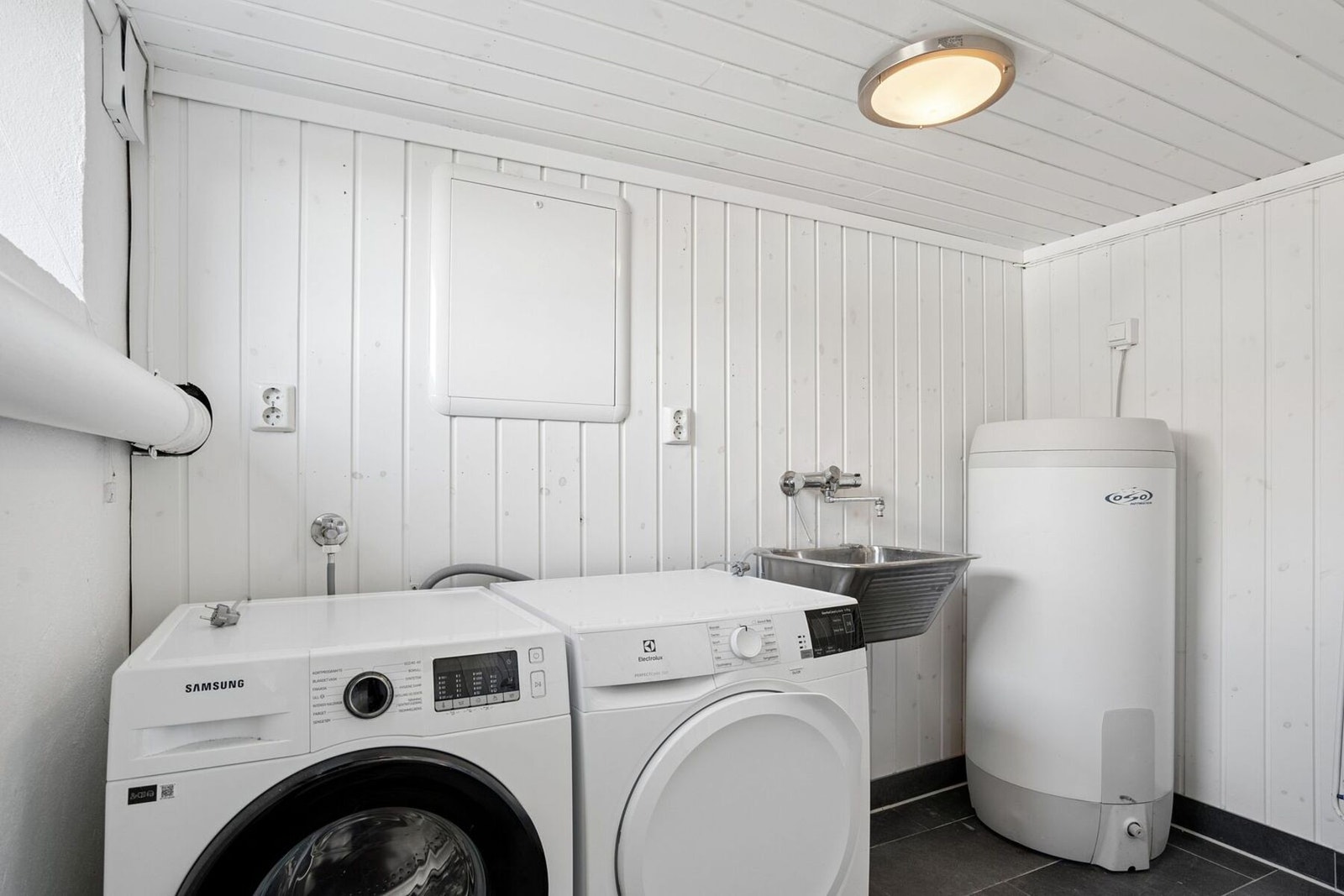 Praktisk vaskerom med opplegg for vaskemaskin, tørketrommel, varmtvannstank, vaskekum og toalett Galleribilde