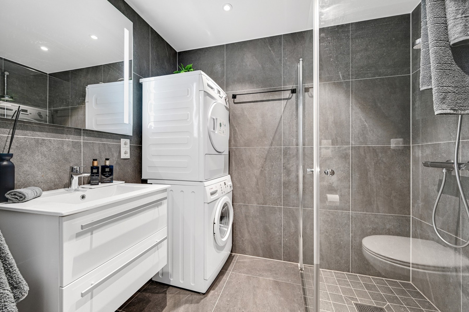 Meget pent flislagt bad med varmekabler innredet med pene dusjvegger, vegghengt klosett, pen baderomsinnredning og opplegg for både vaskemaskin og tørketrommel Galleribilde