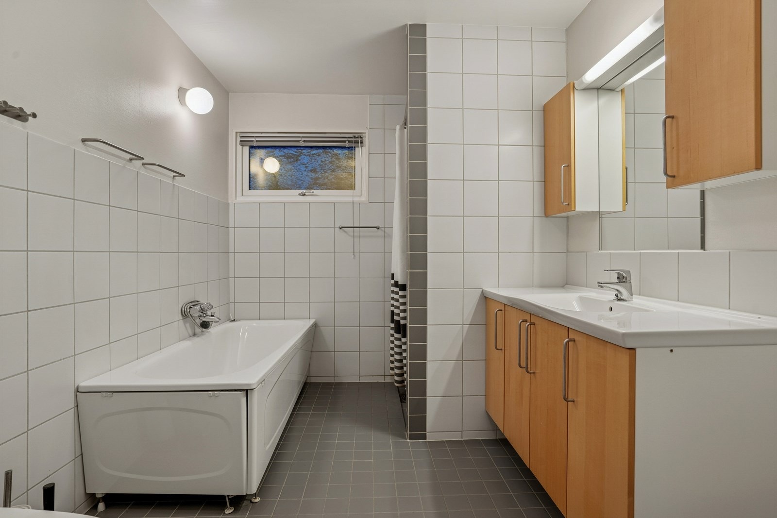 Badet har flislagt gulv og vegger som gir en slitesterk og lettstelt overflate, kombinert med malt tak som skaper en lys og harmonisk atmosfære. Innredningen består av nedfelt servant med tilhørende underskap, toalett, badekar og dusj med hjørnevegger Galleribilde