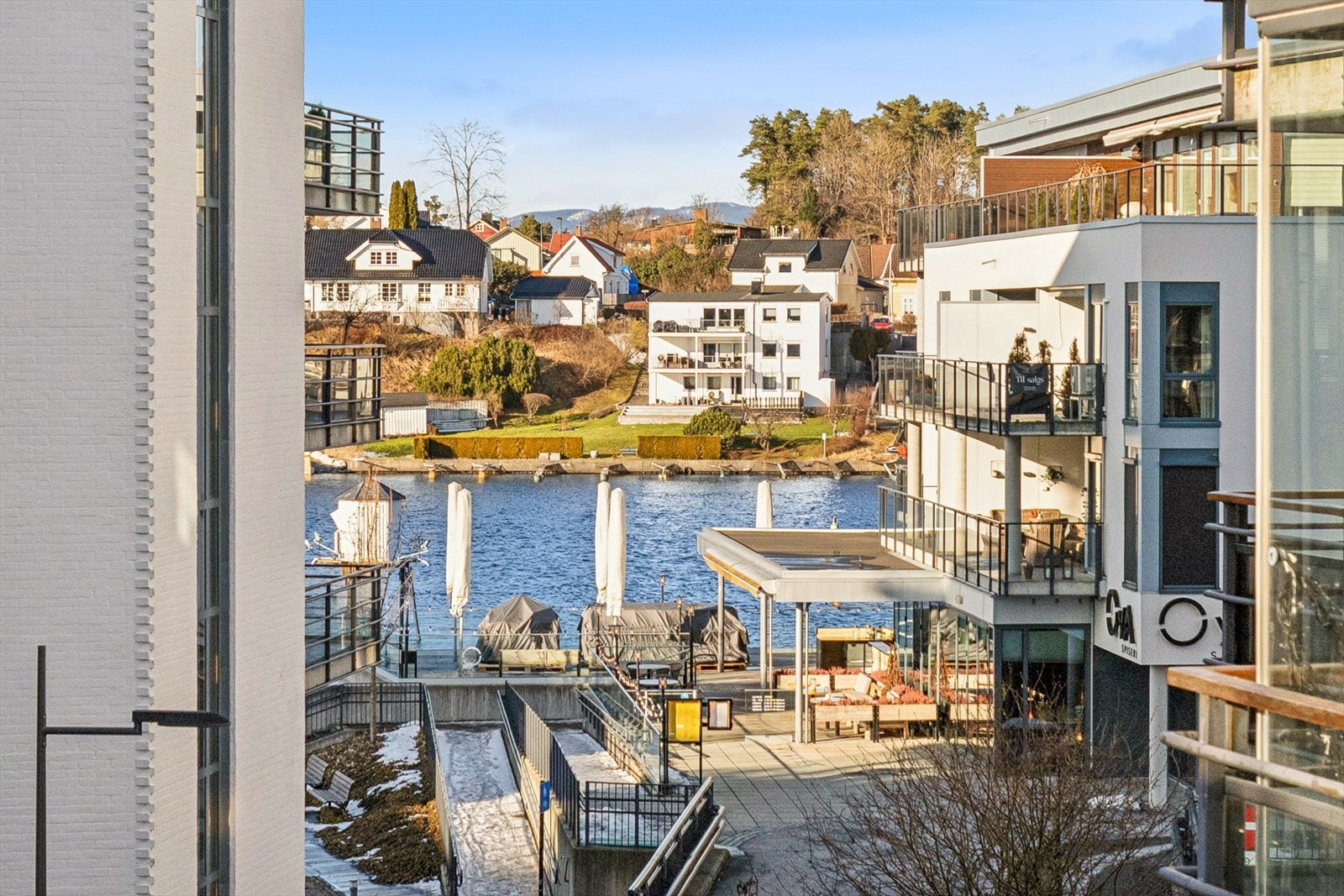 Utsikt mot den idylliske elvepromenaden på Bratsberg Brygge, med glimt av vannet mellom byggene og bebyggelsen på motsatt side av elva. Her får du et levende bymiljø kombinert med den rolige, maritime stemningen som kjennetegner denne delen av Porsgrunn. Galleribilde
