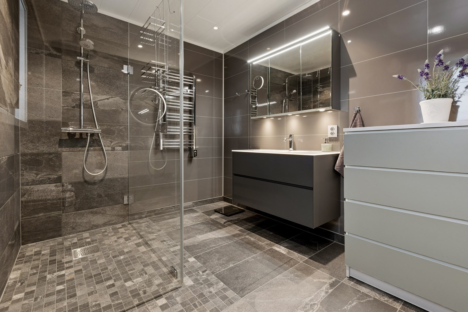 Badet er innredet med vegghengt toalett og servant, speil med integrert belysning og et romslig dusjhjørne med regnfallsdusj. Galleribilde