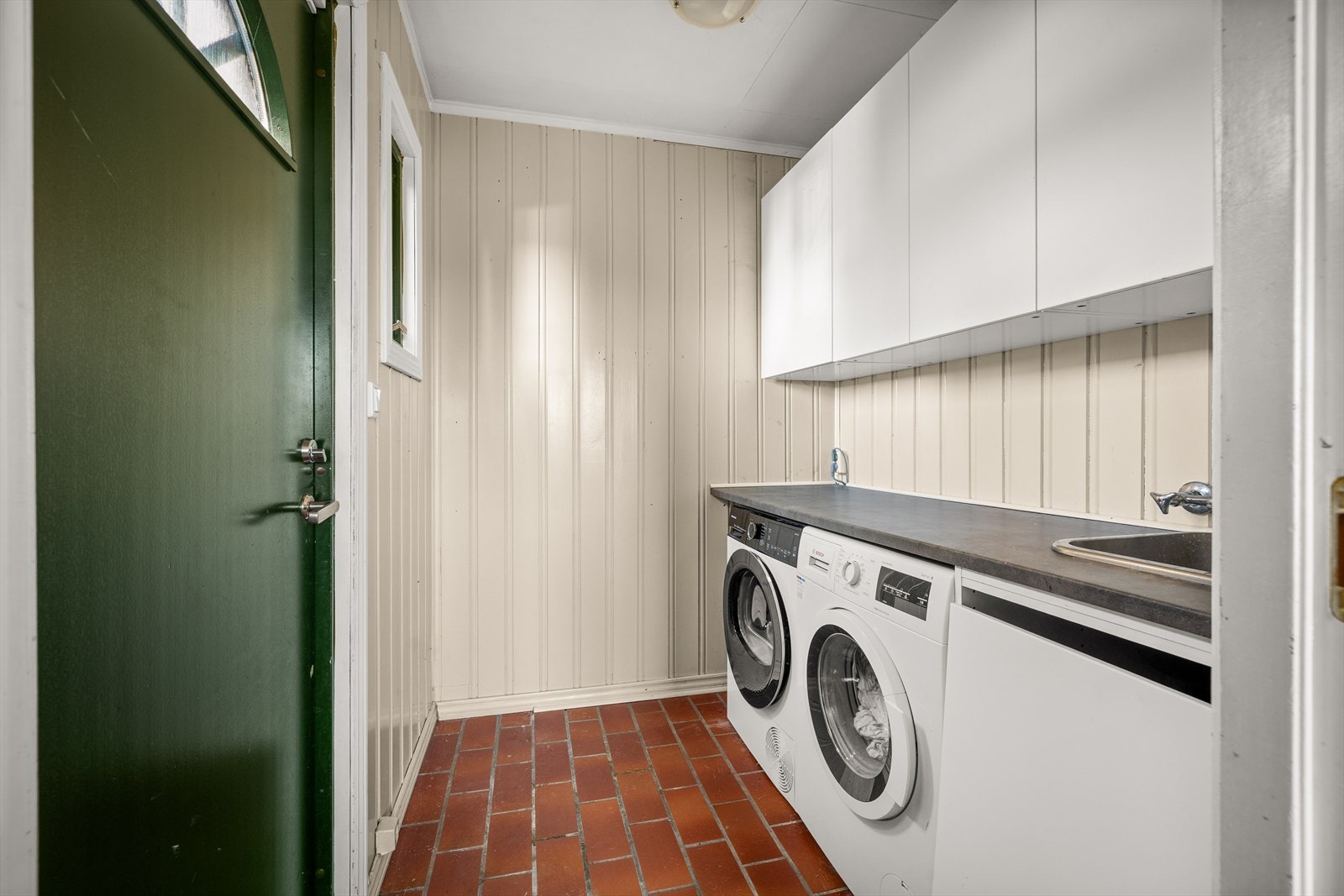 Praktisk vaskerom med flislagt gulv, utslagsvask og opplegg for vaskemaskin. Galleribilde